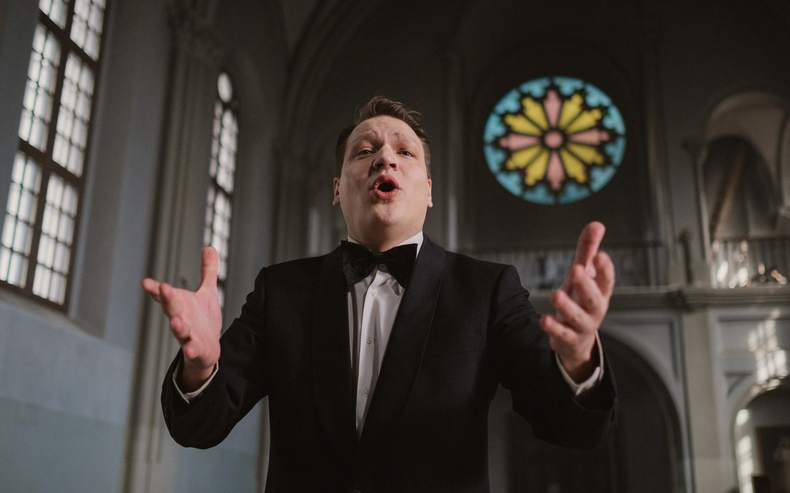 Man in a tuxedo sings expressively beneath a stained-glass rose window in a church