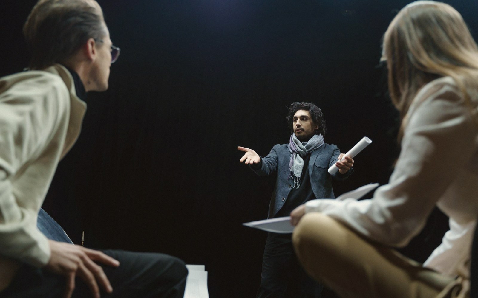 A director gestures while addressing two seated actors during a rehearsal
